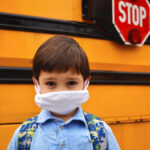 Child with mask on in front of school bus