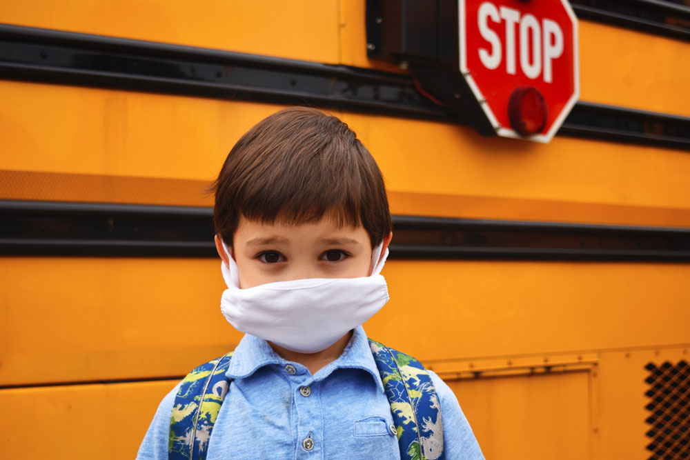 Child with mask on in front of school bus